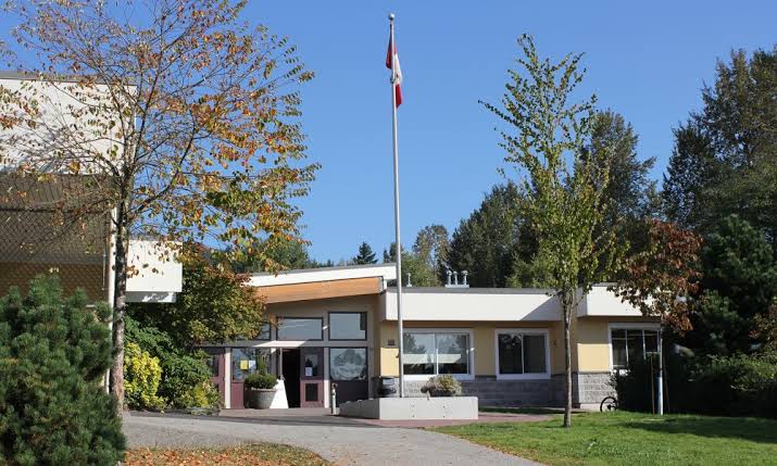 Exterior view of Boundary Elementary School on a sunny day, featuring a single-storey school building with a Canadian flagpole at the entrance, surrounded by trees with early autumn foliage, a grassy lawn, and a paved walkway leading to the front doors.